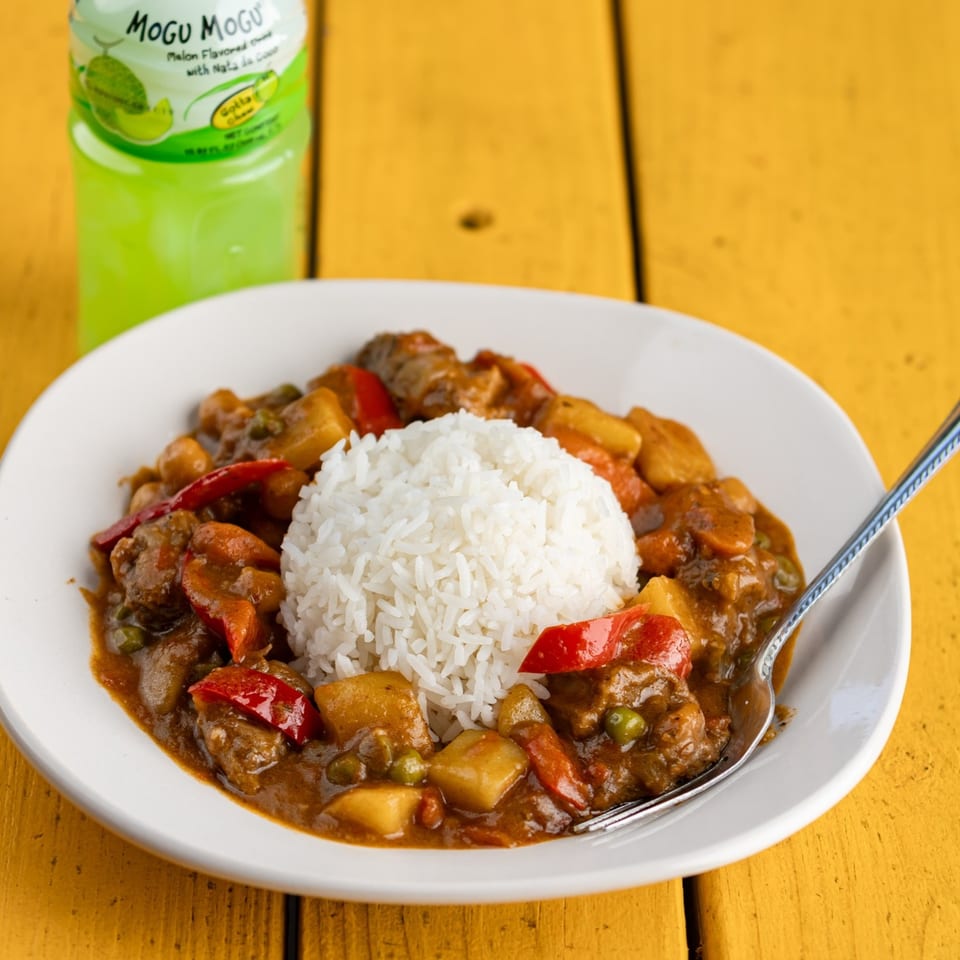Filipino menudo with rice, potatoes, red bell peppers, and a melon drink on a yellow table.