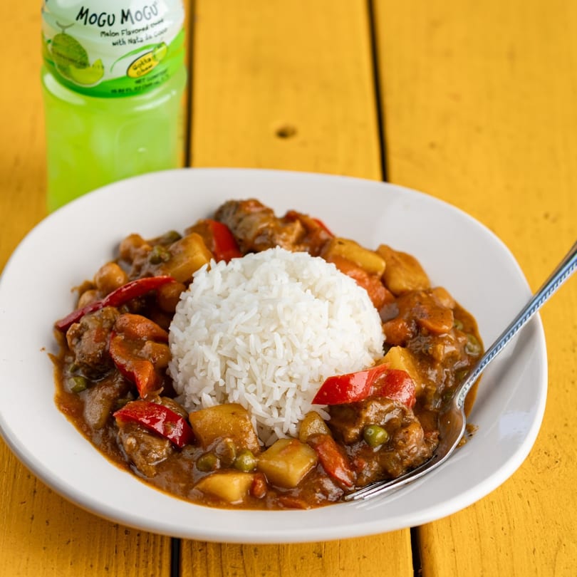 Filipino menudo with rice, potatoes, red bell peppers, and a melon drink on a yellow table.