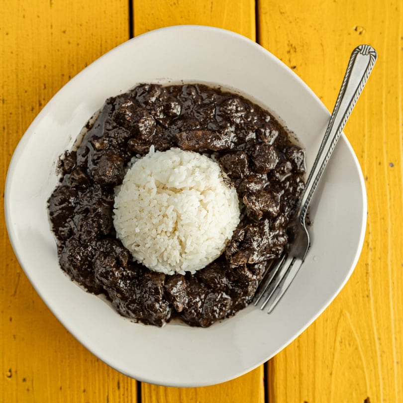 A white plate of Filipino dinuguan, a dark stew, with a scoop of white rice and a fork.